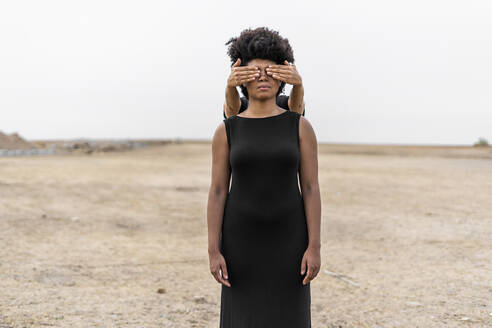 Woman's hands covering eyes of another woman standing in bleak landscape - ERRF01916