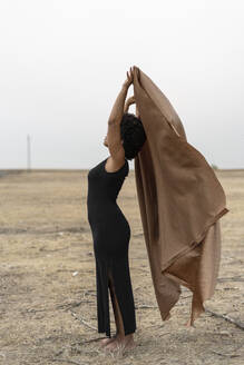 Barfüßige junge Frau in schwarzem Kleid steht in kahler Landschaft und hält eine Decke - ERRF01965