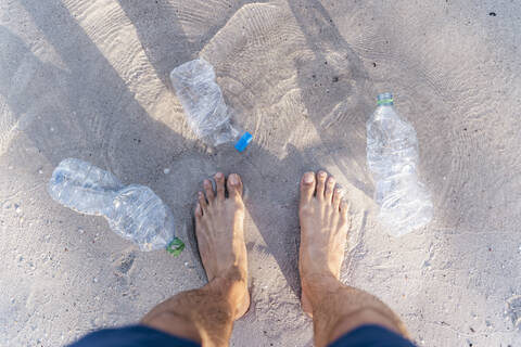 Die Füße eines Mannes am Strand, umgeben von leeren Plastikflaschen, Ansicht von oben, lizenzfreies Stockfoto