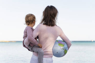 Rückenansicht einer Frau, die ihre kleine Tochter und den Strandball Erde hält und auf den Horizont schaut - DIGF08827