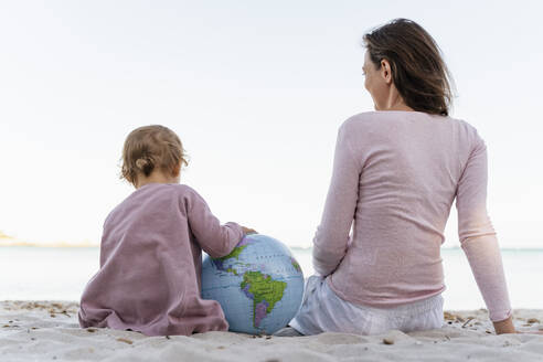 Back view of little girl with Earth beach ball sitting with her mother at seashore - DIGF08829