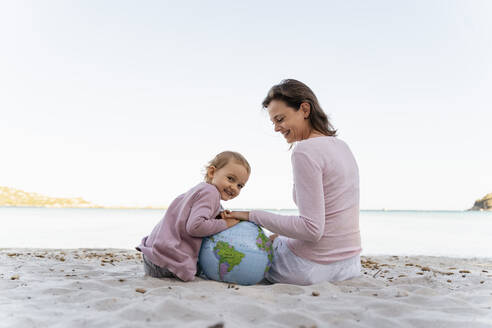 Porträt eines glücklichen kleinen Mädchens, das mit seiner Mutter am Strand sitzt und mit einem Strandball aus Erde spielt - DIGF08830
