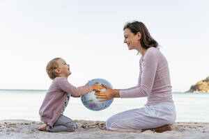 Glückliches kleines Mädchen sitzt mit ihrer Mutter am Strand und spielt mit einem Strandball aus Erde - DIGF08833