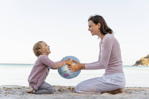 Happy little girl sitting with her mother on the beach playing with Earth beach ball - DIGF08833