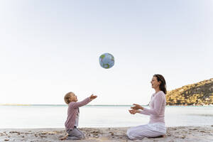 Glückliches kleines Mädchen sitzt mit ihrer Mutter am Strand und spielt mit einem Strandball aus Erde - DIGF08834