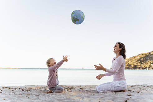 Happy little girl sitting with her mother on the beach playing with Earth beach ball - DIGF08835