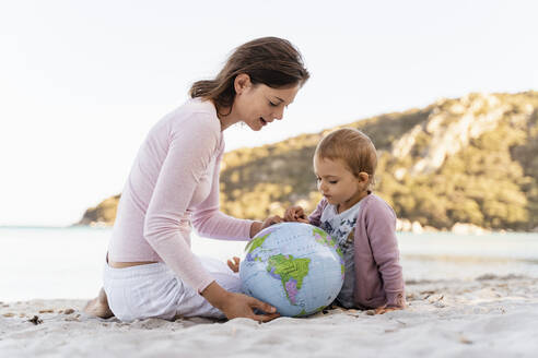 Woman and little daughter sitting on the beach looking together at Earth beach ball - DIGF08843