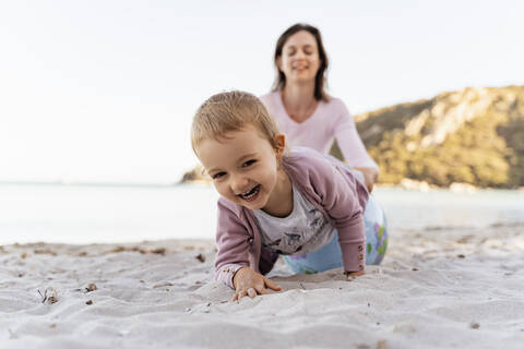 Porträt eines glücklichen kleinen Mädchens, das mit einem Strandball aus Erde spielt, während seine Mutter im Hintergrund sitzt, lizenzfreies Stockfoto