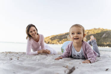 Porträt eines lächelnden kleinen Mädchens am Strand mit einer Mutter, die sie aus dem Hintergrund beobachtet - DIGF08847