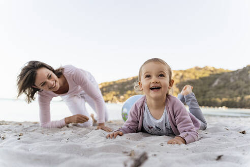 Porträt eines lächelnden glücklichen Mädchens am Strand mit einer Mutter, die sie aus dem Hintergrund beobachtet - DIGF08848