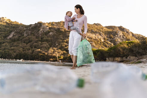 Frau am Strand, die ihre kleine Tochter auf dem Arm trägt und leere Plastikflaschen sammelt - DIGF08857