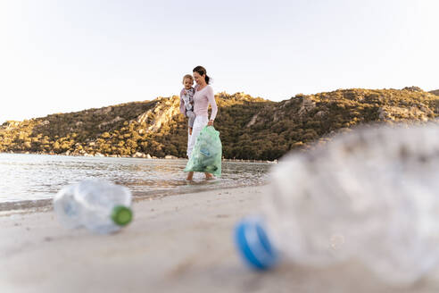 Frau, die ihre kleine Tochter auf dem Arm trägt, sammelt leere Plastikflaschen am Meeresstrand - DIGF08858