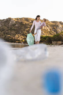 Frau mit kleiner Tochter auf dem Arm sammelt leere Plastikflaschen am Strand - DIGF08861
