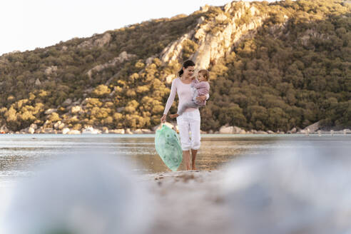 Frau mit kleiner Tochter auf dem Arm sammelt leere Plastikflaschen am Strand - DIGF08862