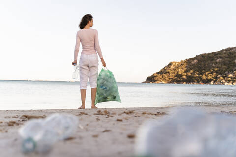 Back view of woman standing on the beach with bin bag of collected empty plastic bottles stock photo