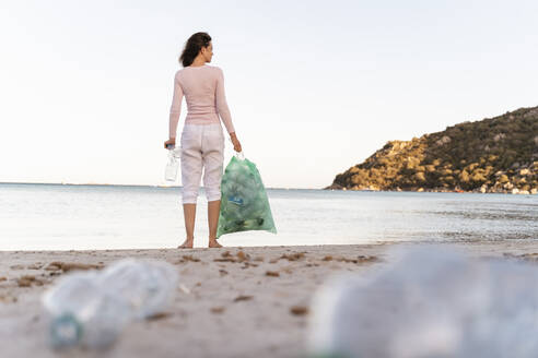 Rückenansicht einer am Strand stehenden Frau mit einem Müllsack mit gesammelten leeren Plastikflaschen - DIGF08864