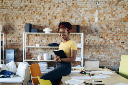 Portrait of confident young woman at table in office holding notebook - SODF00115