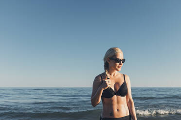 Nachdenkliche Frau mit Bikini und Sonnenbrille gegen Meer und klaren Himmel - CAVF68236