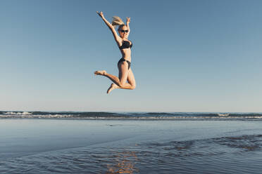 Carefree woman jumping at beach against clear sky - CAVF68237