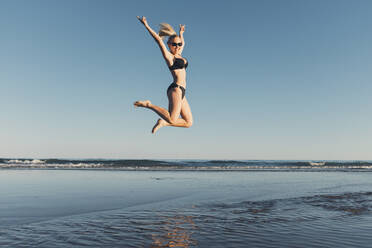 Unbekümmerte Frau springt am Strand gegen den klaren Himmel - CAVF68237