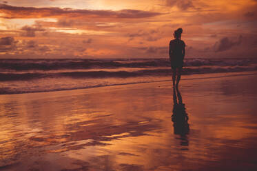 Rear view of woman standing at beach against dramatic sky during sunset - CAVF68256