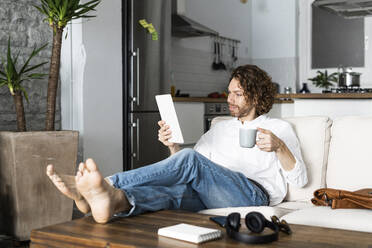 Entspannter Mann sitzt zu Hause auf der Couch und benutzt ein Tablet - GIOF07474