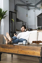 Entspannter Mann sitzt zu Hause auf der Couch mit Handy und Kopfhörern - GIOF07480