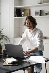 Portrait of confident man sitting at table at home using laptop - GIOF07486