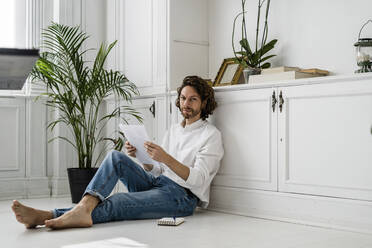 Portrait of man sitting on the floor at home reviewing papers - GIOF07500