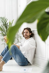 Portrait of man sitting on the floor at home with notepad - GIOF07505