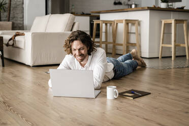 Smiling man lying on the floor at home using laptop - GIOF07513