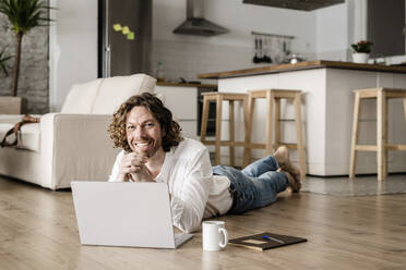 Portrait of smiling man lying on the floor at home using laptop - GIOF07515