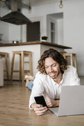 Smiling man lying on the floor at home using cell phone and laptop - GIOF07517