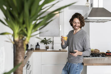 Mann hält Glas Orangensaft und Handy in der Küche zu Hause - GIOF07533