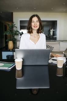 Porträt einer lächelnden Frau, die zu Hause am Tisch mit einem Laptop arbeitet - JRFF03873