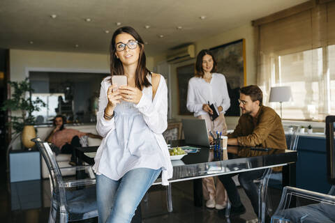 Porträt einer Frau mit Mobiltelefon und Freunden, die im Hintergrund zu Hause arbeiten, lizenzfreies Stockfoto