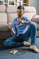 Portrait of young man sitting barefoot on the floor of living room at home looking at smartphone - MGIF00822