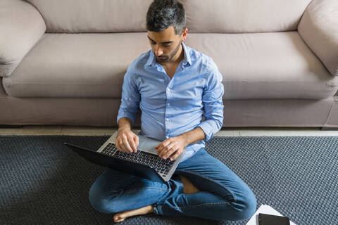 Junger Mann sitzt zu Hause auf dem Boden und benutzt einen Laptop, lizenzfreies Stockfoto