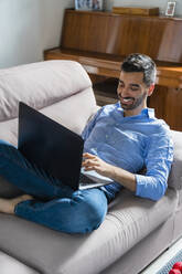 Smiling young man sitting on the couch at home using laptop - MGIF00827