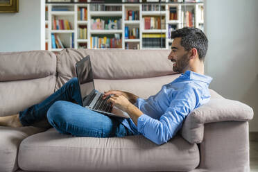 Smiling young man lying on the couch at home using laptop - MGIF00828