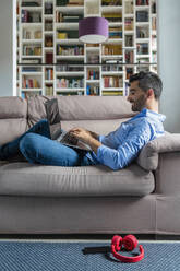 Smiling young man lying on the couch at home using laptop - MGIF00829