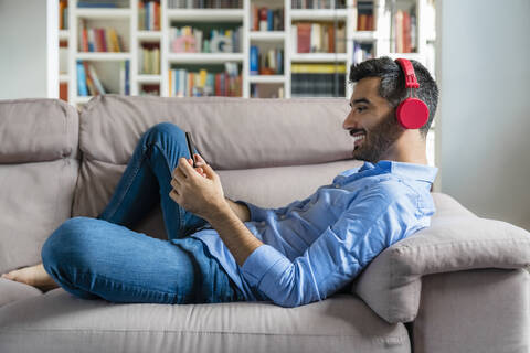 Lächelnder junger Mann, der zu Hause auf der Couch liegt und ein Smartphone und drahtlose Kopfhörer benutzt, lizenzfreies Stockfoto