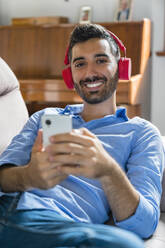 Porträt eines lächelnden jungen Mannes, der zu Hause auf der Couch liegt und ein Smartphone und drahtlose Kopfhörer benutzt - MGIF00833