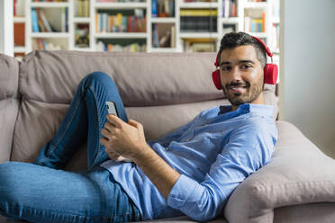 Porträt eines lächelnden jungen Mannes, der zu Hause auf der Couch liegt und ein Smartphone und drahtlose Kopfhörer benutzt - MGIF00834
