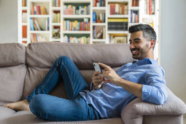 Smiling young man lying on the couch at home using smartphone - MGIF00835
