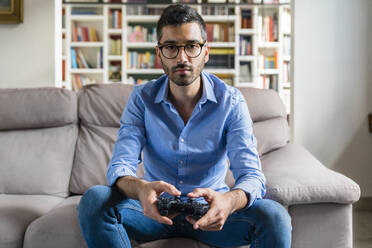 Portrait of serious young man sitting on the couch at home playing video game - MGIF00840