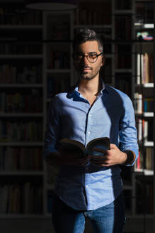 Porträt eines jungen Mannes mit Buch, der zu Hause vor einem Bücherregal steht - MGIF00842