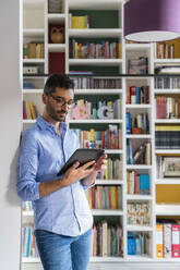 Porträt eines jungen Mannes, der zu Hause vor einem Bücherregal steht und ein digitales Tablet benutzt - MGIF00845