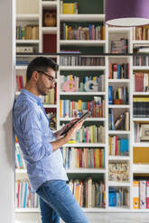 Junger Mann steht zu Hause vor einem Bücherregal und benutzt ein digitales Tablet - MGIF00846