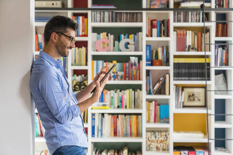 Smiling young man standing in front of bookshelves at home using digital tablet stock photo